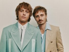 Two men, dressed in suits. standing against a plain background.