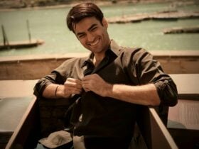 A smiling man in a dark button-down shirt with a sun-drenched harbor and docks in the background