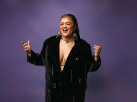 A person with black braided hair and bold earrings stands against a purple background.