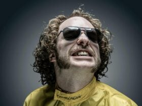 Young man wearing dark aviator glasses with chin length curly hair smiling, wearing a yellow parka.