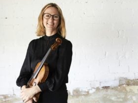 Sophie Rowell standing in front of a rustic white brick wall holding her violin.