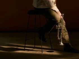 A cropped photo of a trumpet player sitting down on a stool, holding the trumpet in a relaxed way.