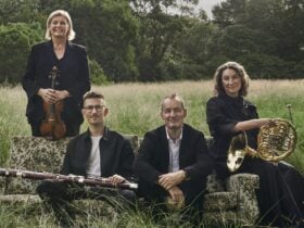 A group of four musicians poses in a grassy field, with two women standing and two men seated.