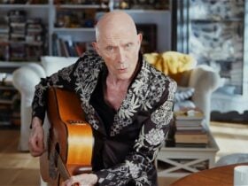 An older person sitting with a guitar in their arms, a living room and bookshelves in the background
