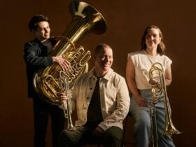 A photo of three ASO brass players holding their instruments and smiling in a studio setting.
