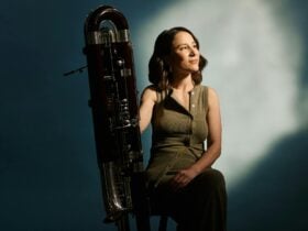 A photo of contra bassoon player holding her instrument, seated, in a studio setting.