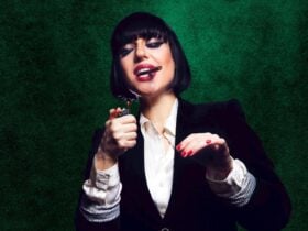 A woman with a brunette bob, red lips and a black suit jacket lights up a cigar
