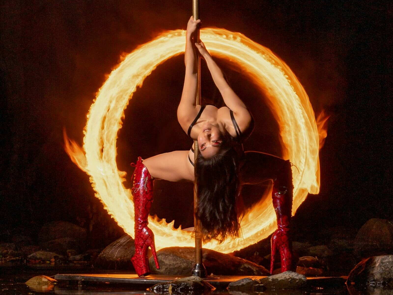 long hair Asian woman red knee high stiletto boots back bend on pole surrounded by ring of fire