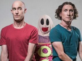 Two men sit side-by-side with deadpan expressions, flanking a tall puppet with red mouth