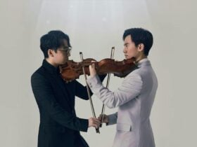 Two men wearing suits stand facing each other, each holding a violin.