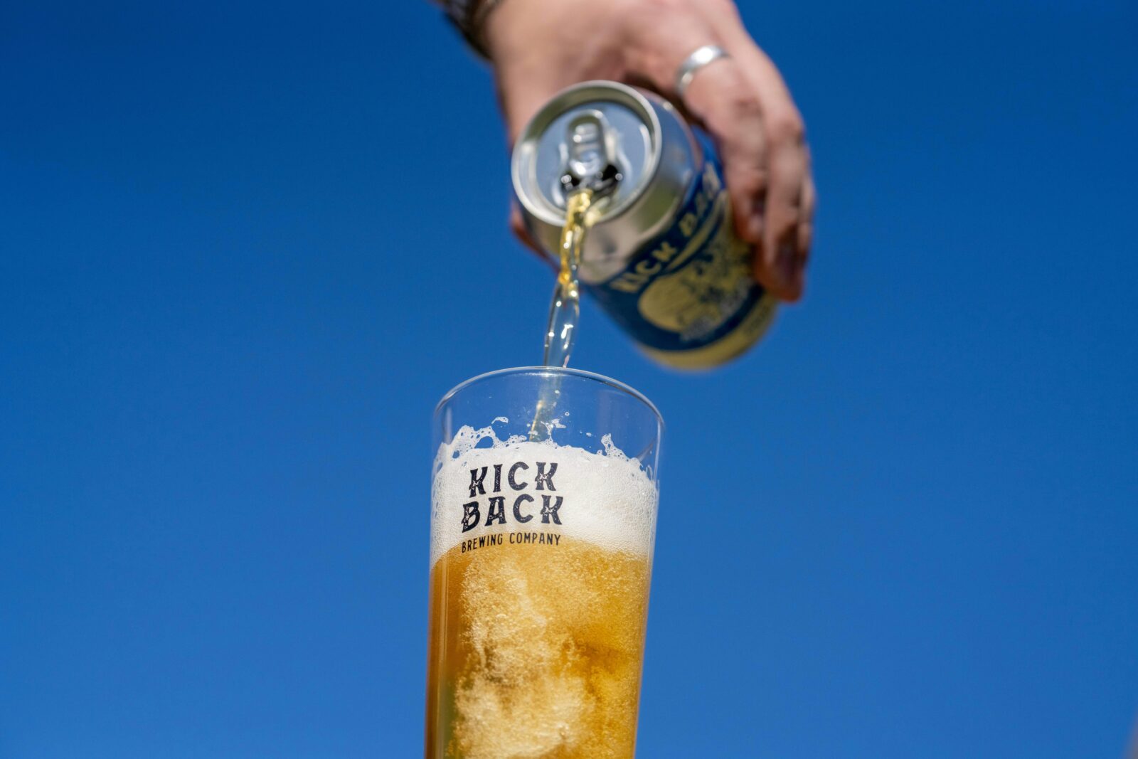 Hand pouring a can of Kick Back Brewing beer into a branded glass with blue sky backdrop.
