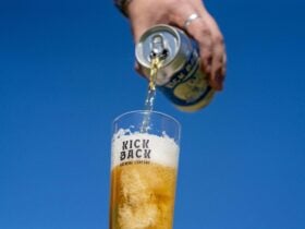 Hand pouring a can of Kick Back Brewing beer into a branded glass with blue sky backdrop.
