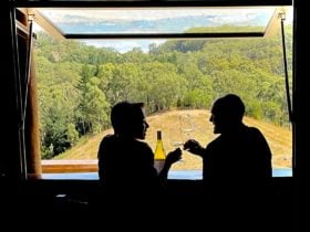 a couple sit in front of a view of Australian bushland drinking a glass of wine