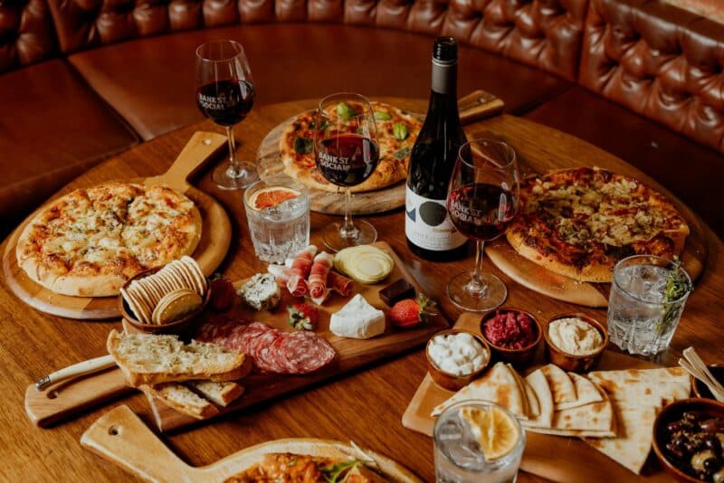 Wooden table filled with delicious pizza, a cheese board in the cenre and wine.