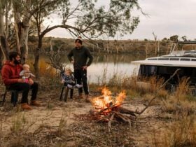 Family by a campfire beside a houseboat on the Murray River, surrounded by trees and calm water.