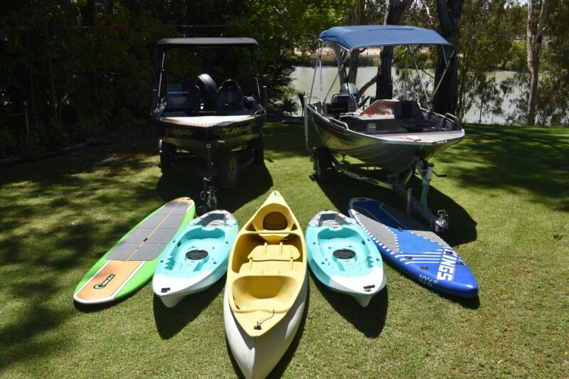 Two hire boats, kayaks and stand-up paddleboards set up on lawn beside the Murray River in Waikerie