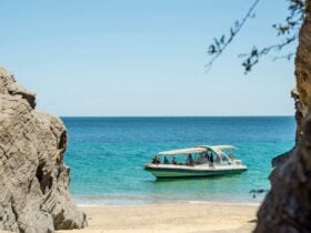 A boat with passengers anchored near a secluded beach, framed by rocky cliffs and clear blue water.