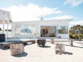 Exterior view of a white beach house with an open patio area, fire pit, and rustic wooden seating