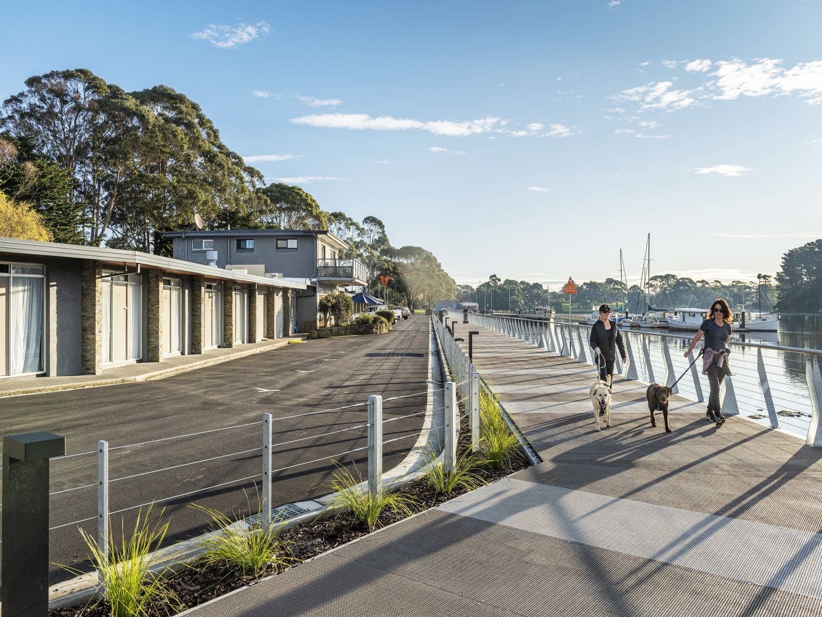 Two people walking their dogs along a modern boardwalk along the Inglis river motel on left