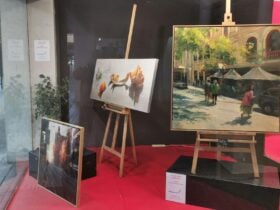 Paintings displayed in a gallery window, including realist still life and street scene artworks on e