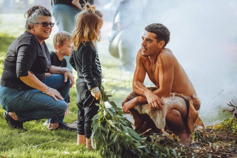 A Palawa man crouching down talking to a young child while a ceremonial fire burns in the background