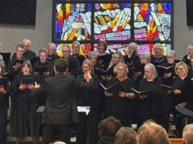 A group of singers dressed in black sing in front of a stained glass window.
