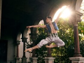 Ballet dancer leaps in ornate costume beneath stone arches, sunlight behind.