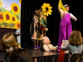 A woman in pink overalls performs on a stage with toys while children watch