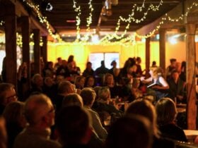 Darkly lit audience watching a performance, with warm string lights hanging overhead.