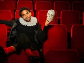 A man in a ruff holding a skull sitting in red theatre seats