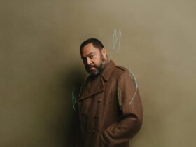 dark skin man in brown jacked standing in front of light brown wall