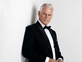 Michael Cormick in a tux leaning on a white wall smouldering