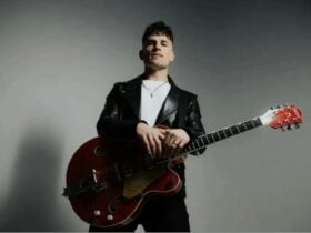 The image is a low-angle, high-contrast portrait of a musician against a neutral grey background. Th