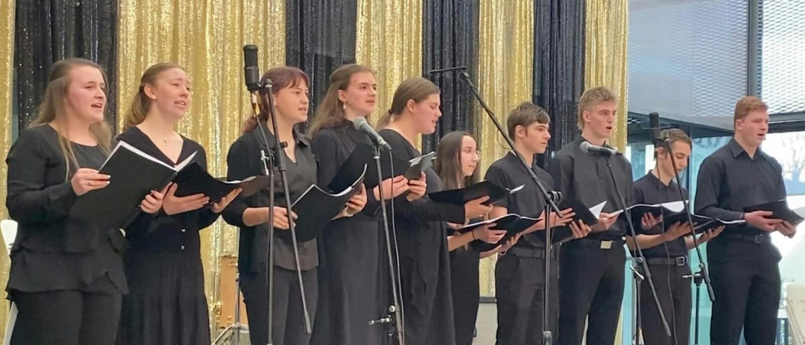 A group of singers holding books wearing black on a stage in front of stand microphones