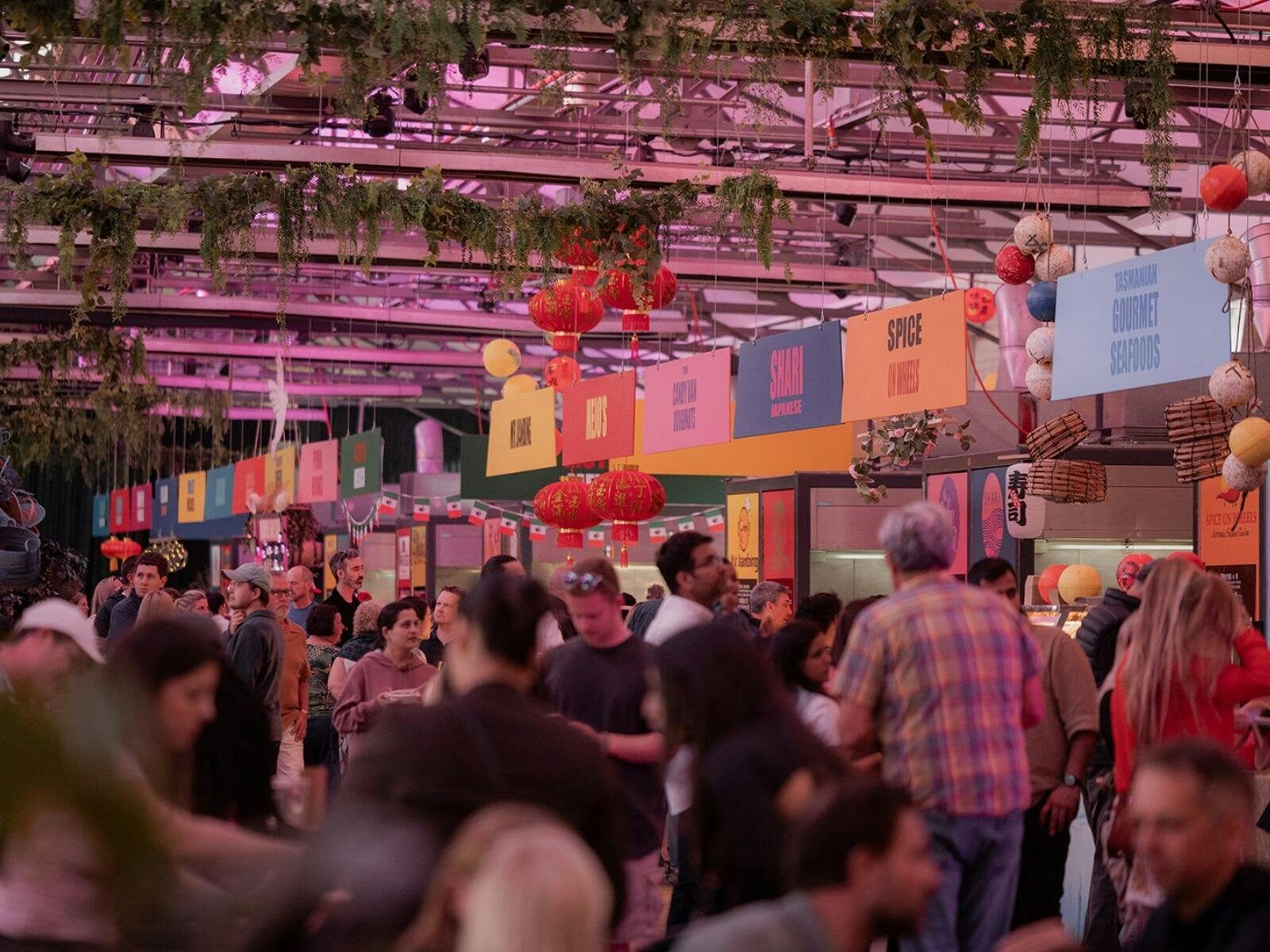 Evening crowds enjoy food stalls beneath lanterns and greenery at a vibrant festival.