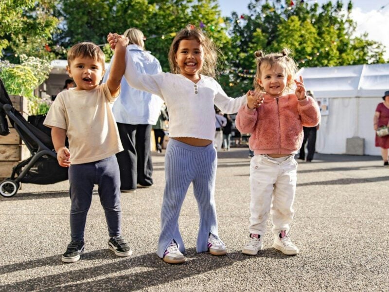 Three smiling children hold hands outdoors at a sunny, family-friendly festival event.
