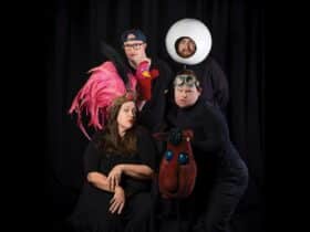 Four performers dressed in black on a black background. Each one folding or wearing a colourful prop