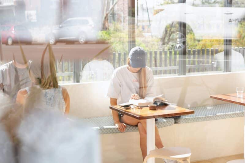 A woman reads a book in the sunshine at a table inside Abercrombie Coffee.