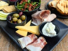 Some charcutterie, cheeses and accompaniments on a black plate with toasted bread on the side