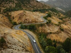 A vehicle drives along a winding road toward a mountain town in the distance