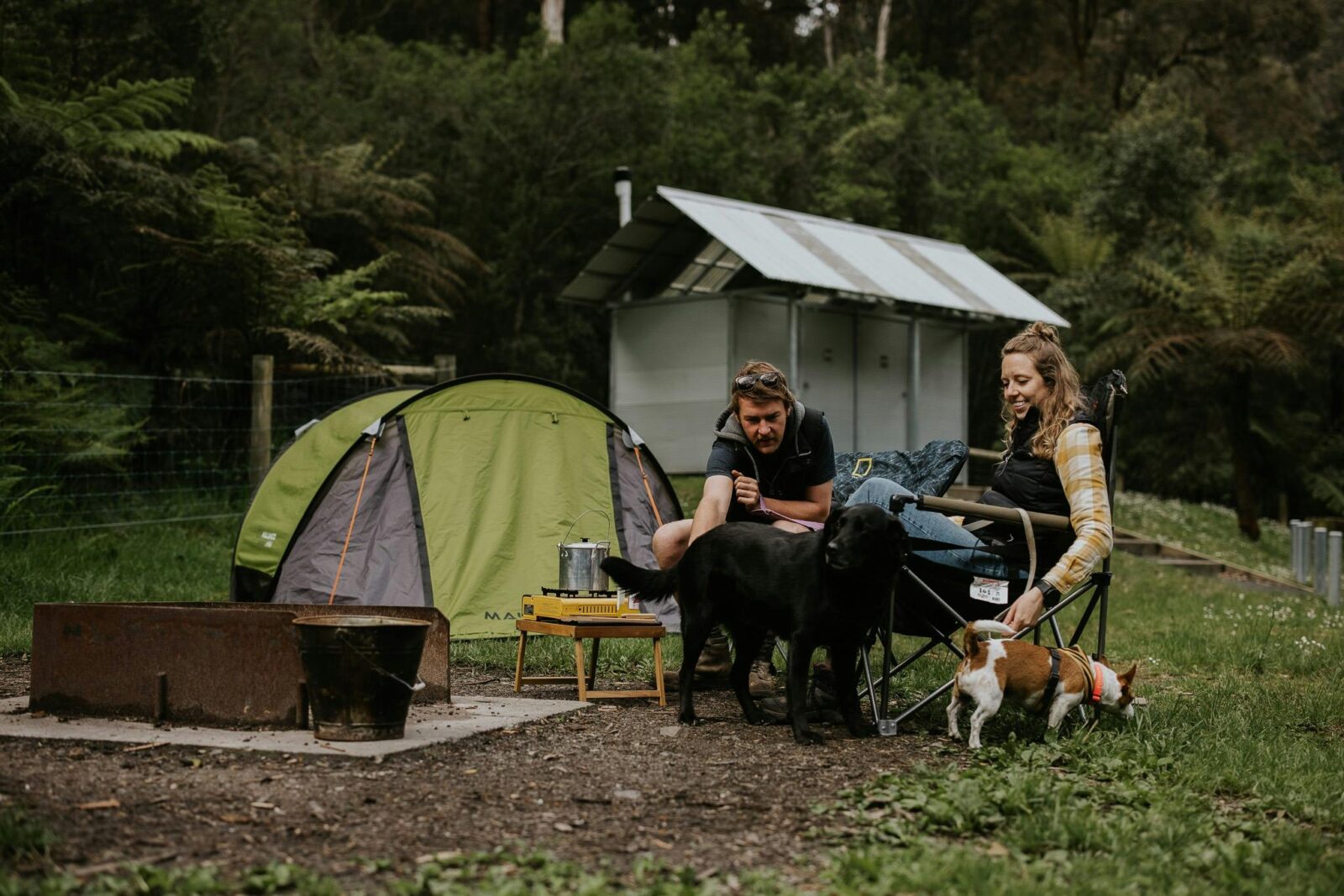 Two people and a dog in front of a campfire BBQ and tent