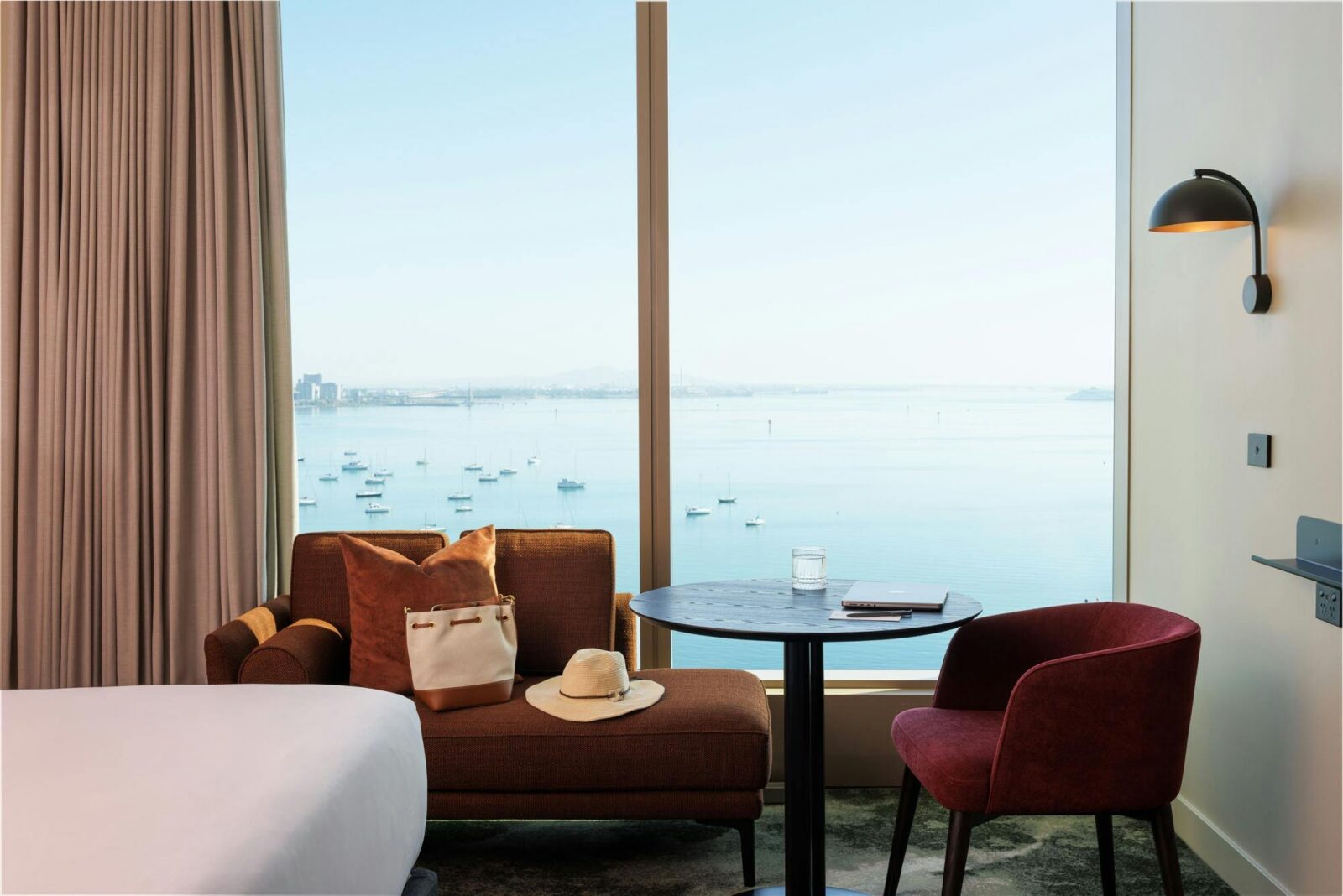 A brown couch with a hat and a bag are in front of floor to ceiling windows overlooking the bay