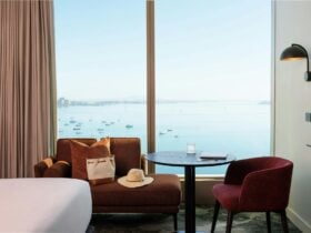 A brown couch with a hat and a bag are in front of floor to ceiling windows overlooking the bay