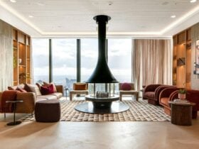 A large room with central fire pit, floor to ceiling windows and orange and brown furniture/decor..