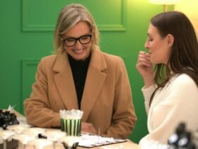 Two women blending fragrance oils at a workshop table with rows of small black bottles in front of t