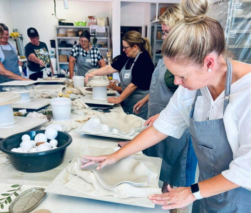 An image of a group of women standing around a large work bench, making pottery