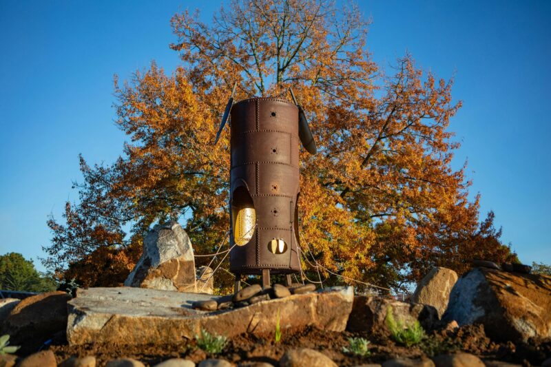 A tall metal sculpture sits atop a boulder infront of a large autumnal tree and blue sky.