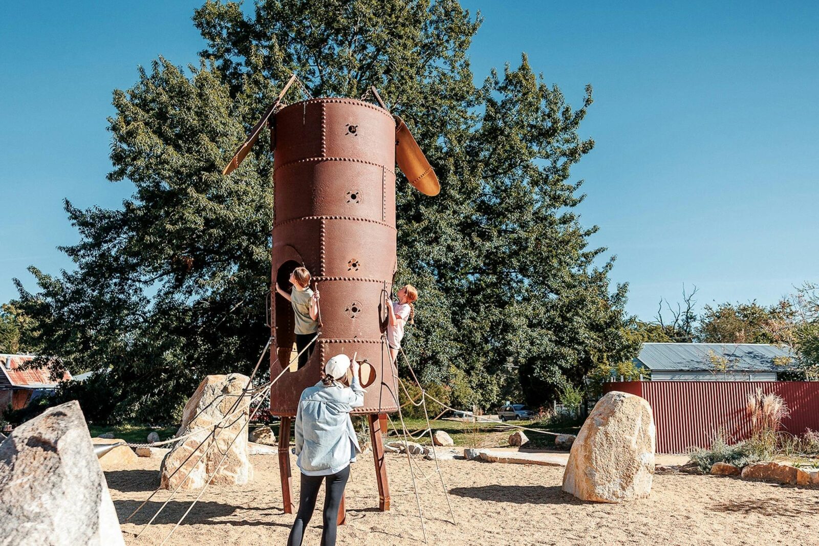 A woman watches as children play on a tall metal tower surrounded by ropes and boulders