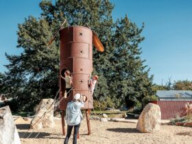 A woman watches as children play on a tall metal tower surrounded by ropes and boulders