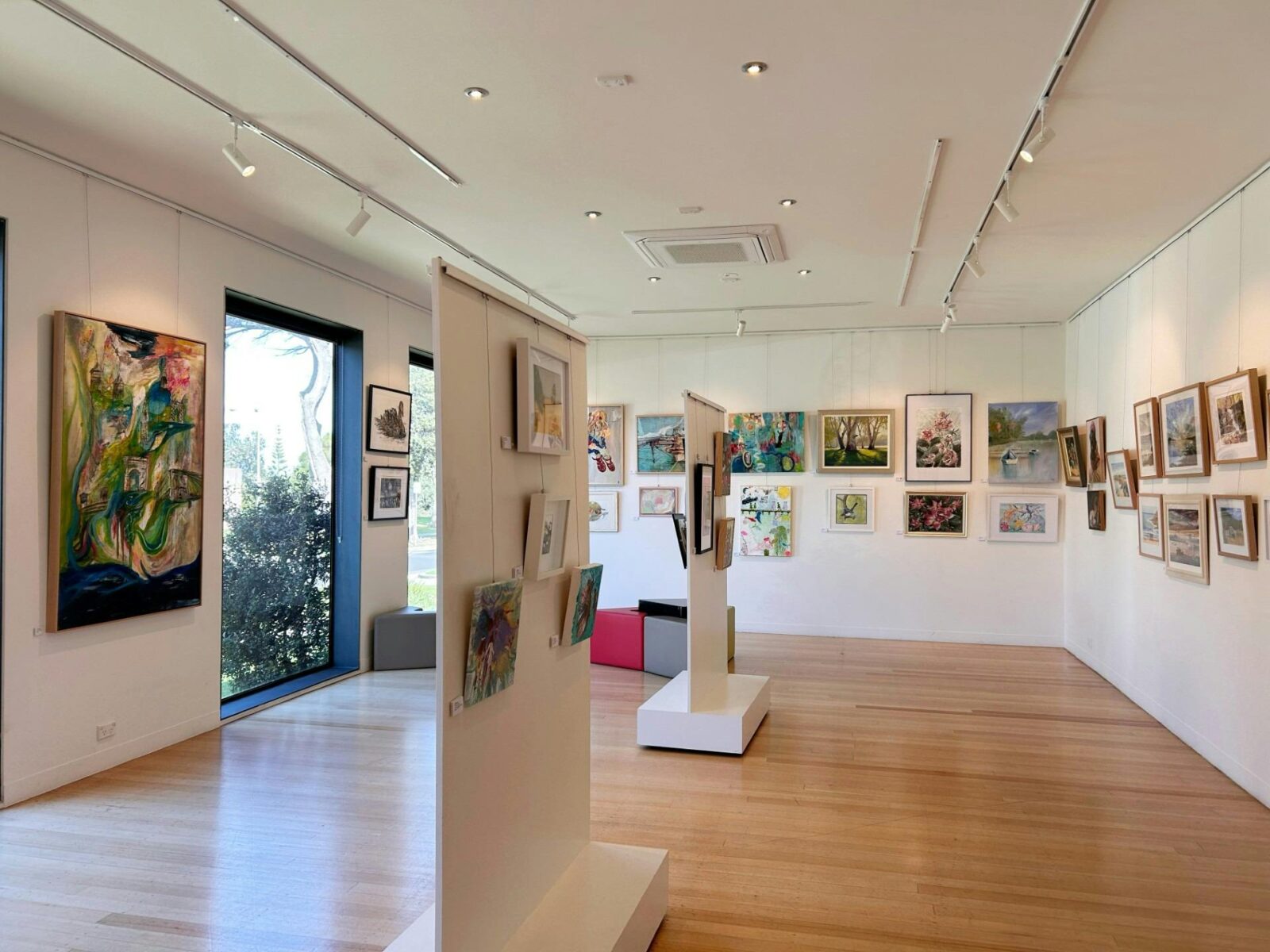 Multiple artworks of different mediums, hanging in a light-filled gallery space with large windows